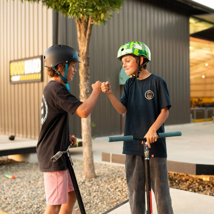 two kids with scooters wearing helmets in front of a warehouse fist bumping each other 