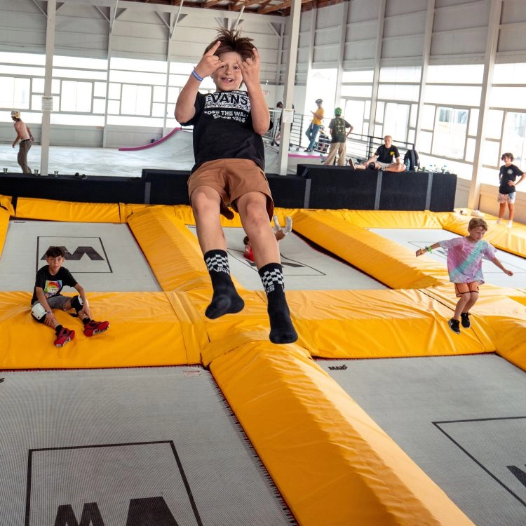 kids jumping on trampolines in a trampoline park, one kid at center of photo is high in the air doing horn symbols with his hands